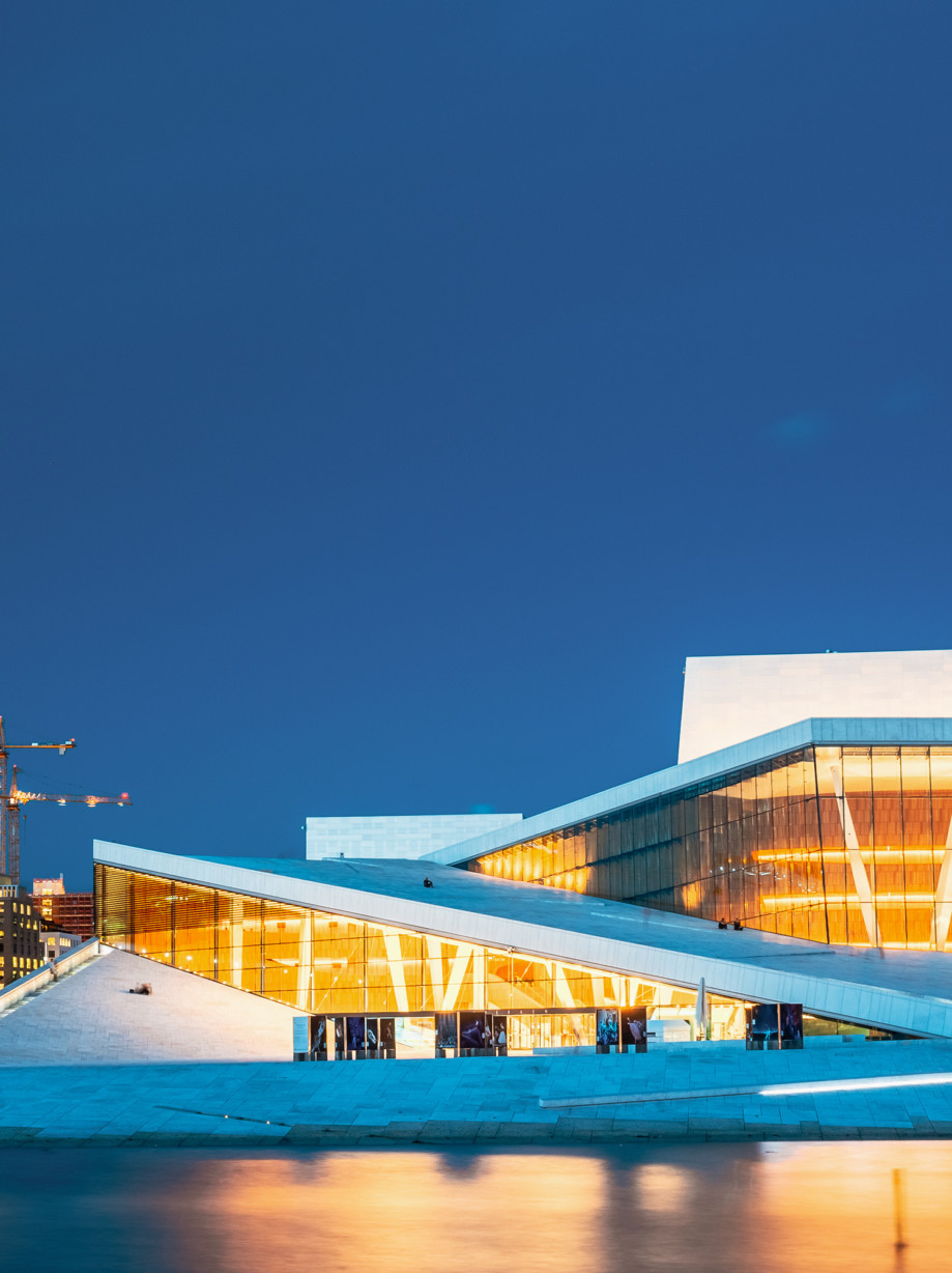 Oslo Norway. Evening View Of Illuminated Opera Ballet House Among High-Rise Buildings Under Blue Sky